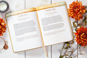 Open book with text on a wooden surface with orange flowers and a candle.
