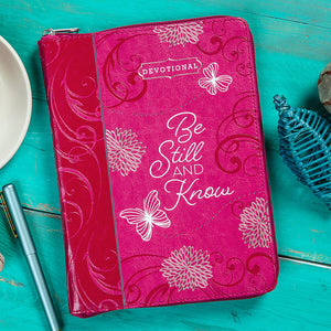 Pink devotional book with floral designs and 'Be Still and Know' text on a teal surface.