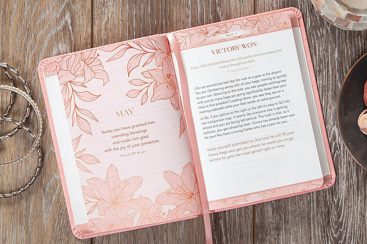 An open pink floral journal or prayer book on a rustic wooden surface, with printed text on the right page and decorative pink illustrations on both pages. A chain and a small dish are partially visible at the edges.