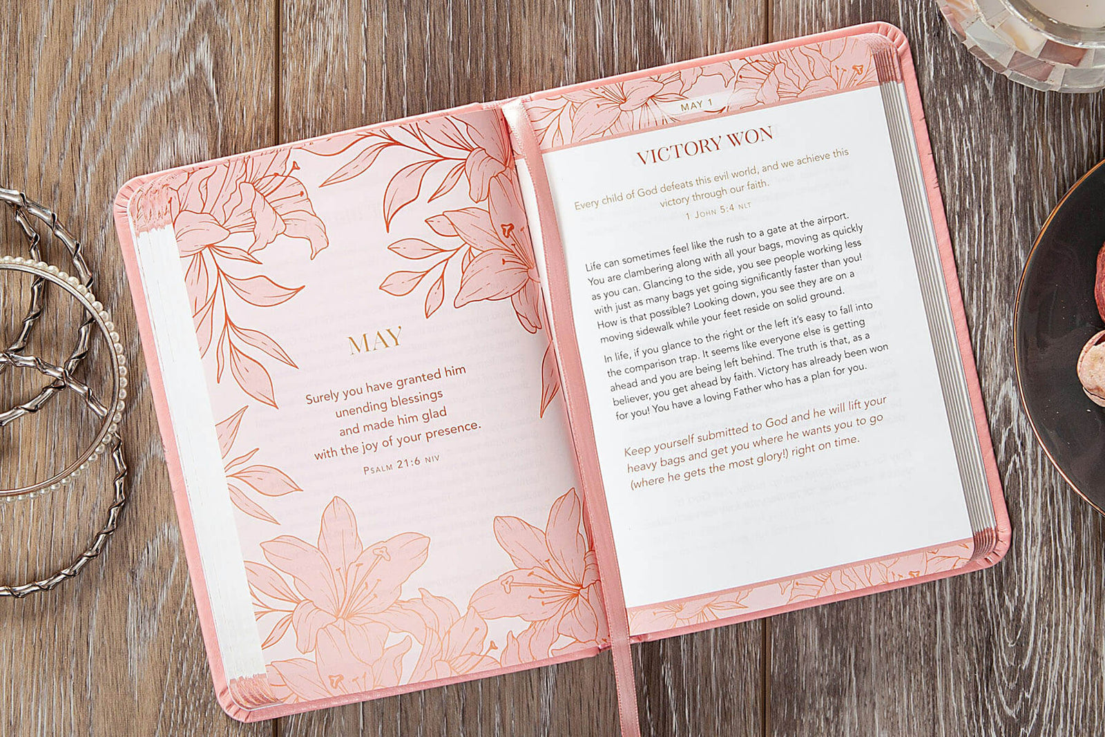A pink hardcover devotional book titled 'Consider the Lilies: 365 Devotions for Women' lies on a rustic wooden surface, surrounded by decorative silver rings and a glass candle holder.