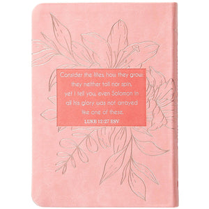 A pink hardcover notebook featuring embossed floral line art and a central coral-colored text block with a Bible verse from Luke 12:27 ESV.