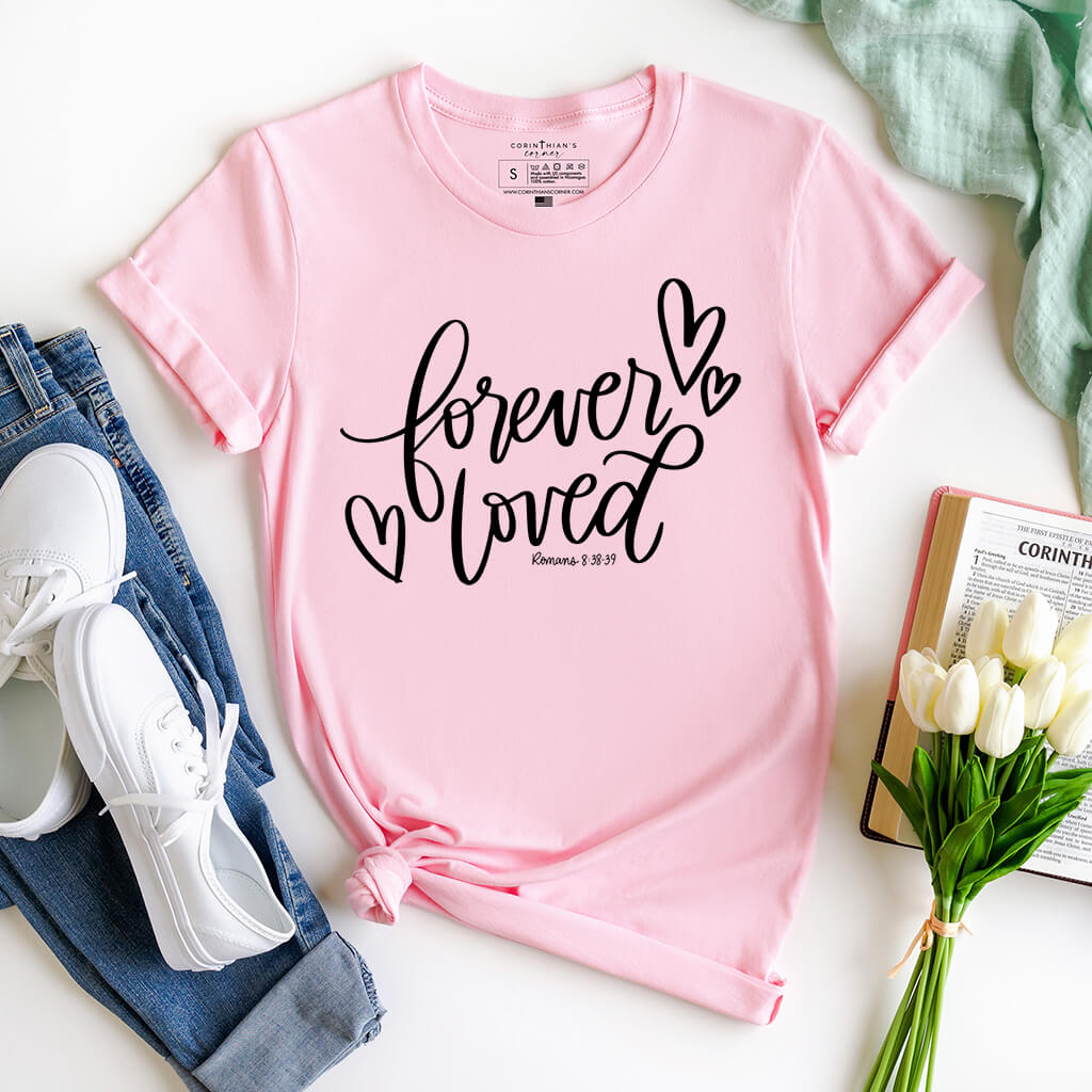 A soft pink t-shirt with the phrase Forever Loved - Romans 8:38-39 printed in elegant white black across the front
