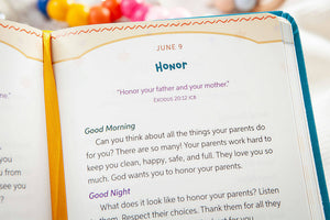 An open Bible or prayer journal showing a printed page with the heading 'HONOR' and a scripture reference, photographed close up with a colorful ribbon bookmark and blurred decorative items in the background.