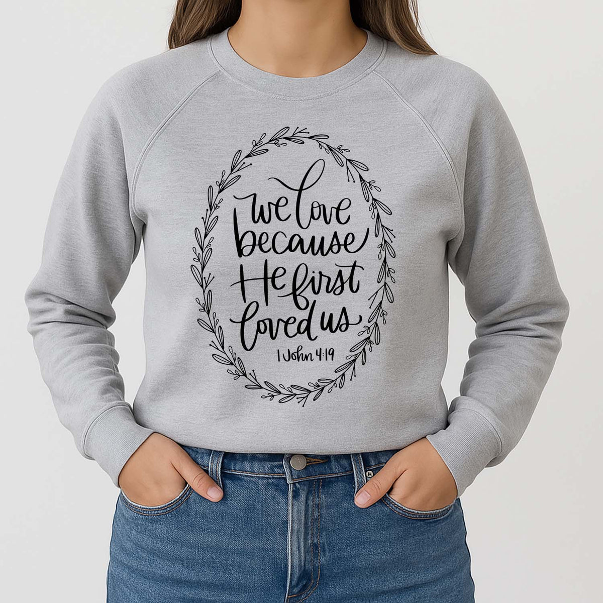 Gray sweatshirt with 1 John 4:19 inscription and floral design worn by a woman in blue jeans