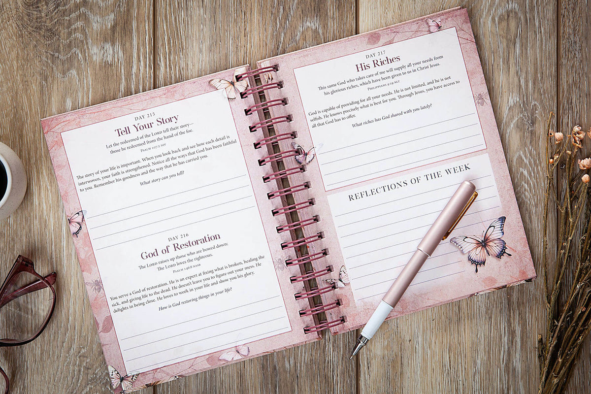 An open spiral-bound journal with pink floral pages laid on a wooden surface, alongside a rose-gold pen, eyeglasses, and dried flowers. The pages contain reflective journaling prompts such as 'Tell Your Story,' 'His Words,' and 'Reflections of the Week.