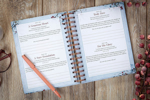 An open spiral-bound journal laid flat on a rustic wooden surface, showing two printed pages with inspirational prompts and quotes. A peach-colored pen rests on the left page, and red berries are placed around the journal as styling props.