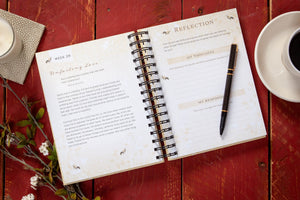 Open spiral-bound notebook with a pen on a red wooden surface with a cup of coffee and decorative elements.