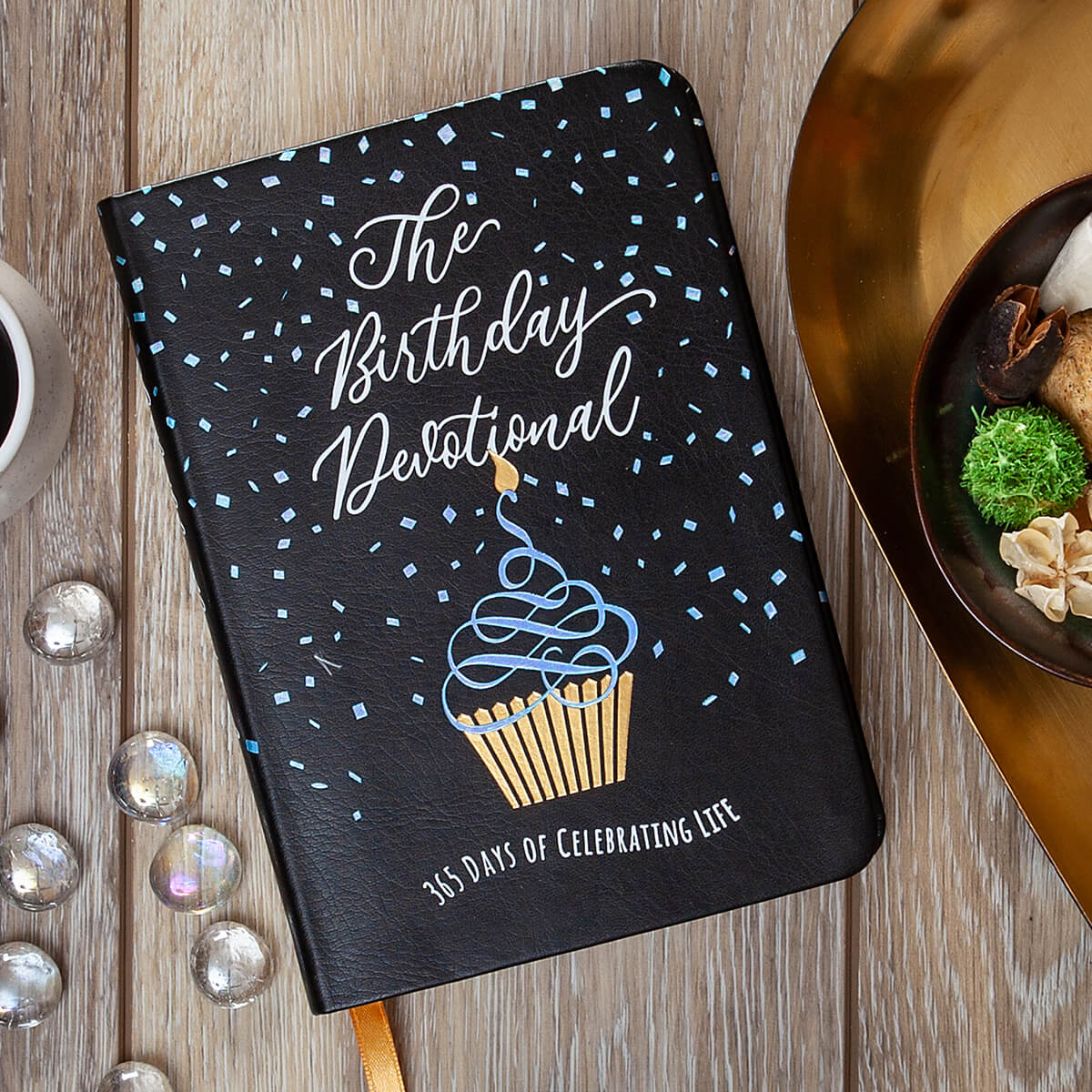 Black notebook titled 'The Birthday Devotional' with a cupcake illustration on a wooden surface.