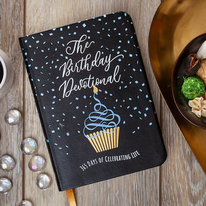 Black notebook titled 'The Birthday Devotional' with a cupcake illustration on a wooden surface.
