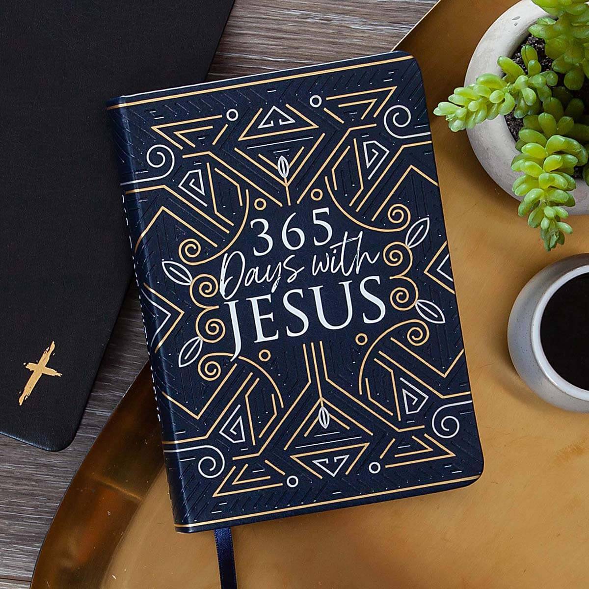 Notebook titled '365 Days with Jesus' on a wooden surface with a plant and coffee cup.
