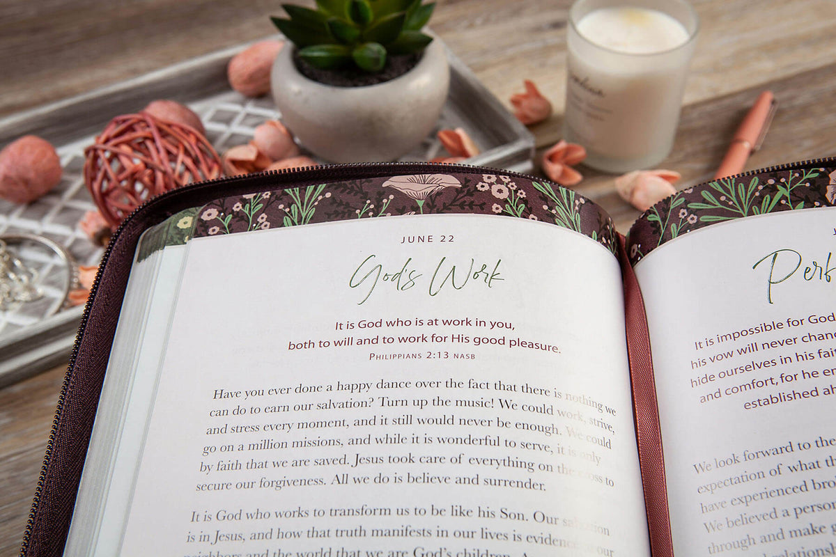 Open book with a decorative cover on a wooden surface with a plant and candle in the background.