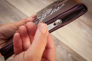 A close-up of a person's hands holding a dark brown leather zippered clutch or wallet, showing the silver zipper pull and the white script logo on the front, set against a blurred light wood background.