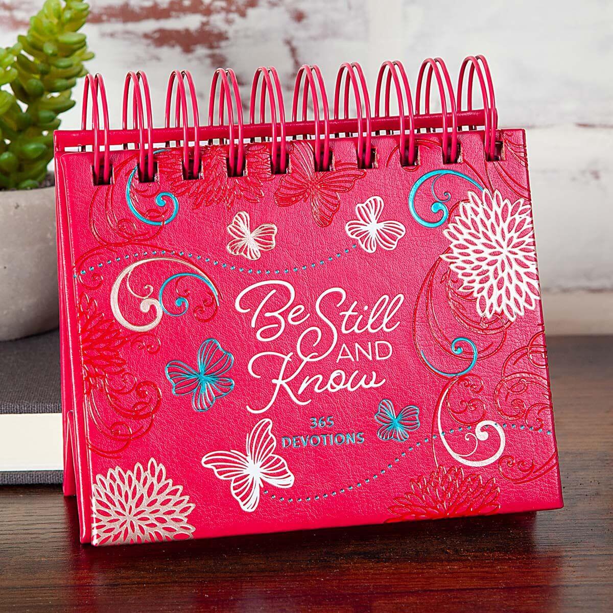 Pink devotional book titled 'Be Still and Know' with floral and butterfly designs on a wooden surface.