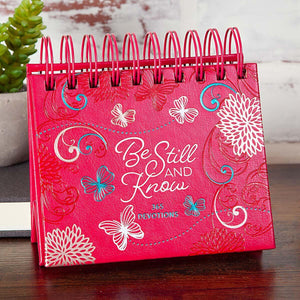 Pink devotional book titled 'Be Still and Know' with floral and butterfly designs on a wooden surface.