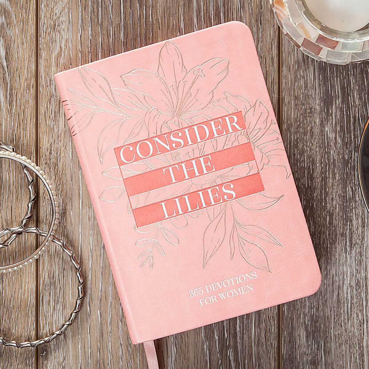 A pink hardcover devotional book titled 'Consider the Lilies: 365 Devotions for Women' lies on a rustic wooden surface, surrounded by decorative silver rings and a glass candle holder.