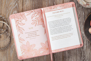 An open pink floral journal or prayer book on a rustic wooden surface, with printed text on the right page and decorative pink illustrations on both pages. A chain and a small dish are partially visible at the edges.