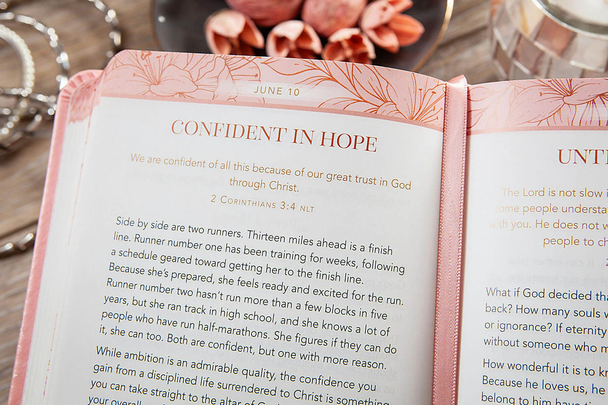 An open Bible with a pink floral cover and white pages, showing the text 'CONFIDENT IN HOPE' and scripture from 2 Corinthians 3:4-5. The book is photographed close-up with a blurred background containing a silver chain and warm-toned objects.