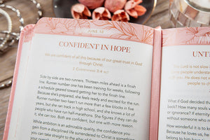 An open Bible with a pink floral cover and white pages, showing the text 'CONFIDENT IN HOPE' and scripture from 2 Corinthians 3:4-5. The book is photographed close-up with a blurred background containing a silver chain and warm-toned objects.