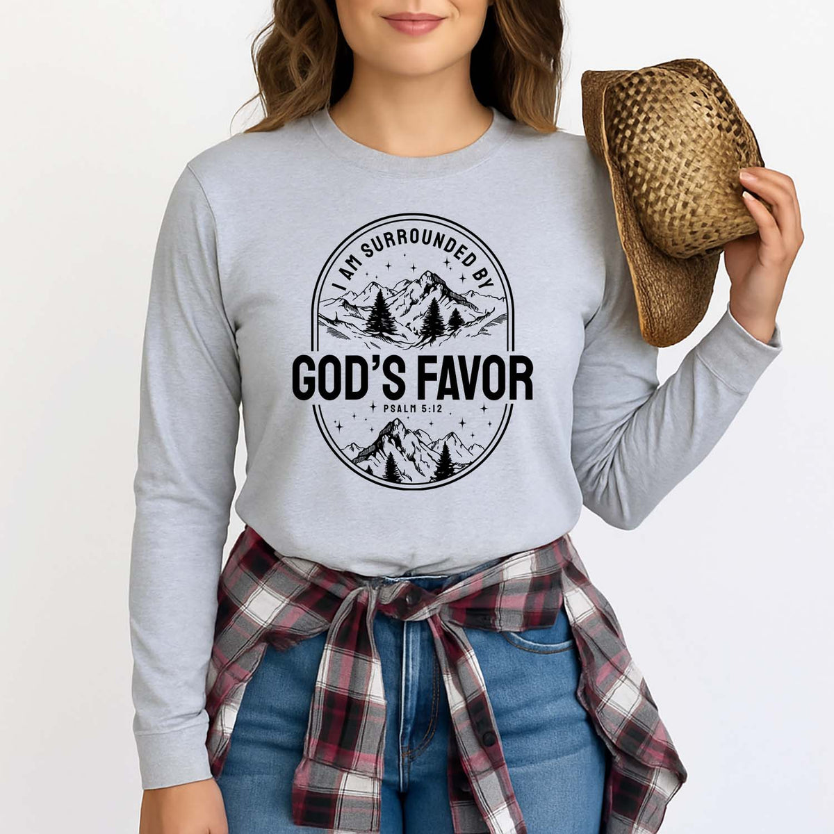 Woman wearing a gray long-sleeve shirt with 'I am surrounded by God's favor' text and mountain graphic, holding a straw hat.