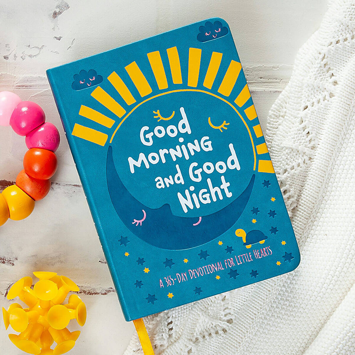 A hardcover children's devotional book titled 'Good Morning and Good Night' lies on a white textured surface next to colorful beads and a yellow flower. The cover is bright blue with yellow sun rays, white lettering, and small cartoon clouds and stars.