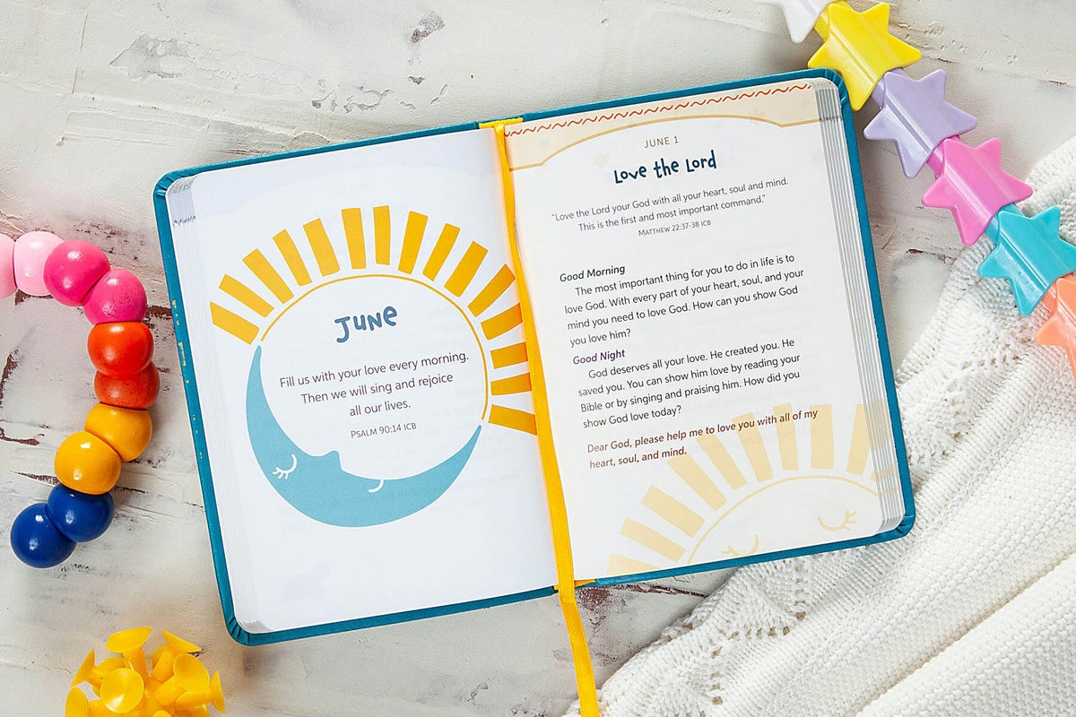 An open Bible with a teal cover and yellow ribbon bookmark, laid flat on a white textured surface with colorful star and bead props around it. The left page shows a June devotional with a sunburst illustration, and the right page contains a morning prayer titled 'Love the Lord'.