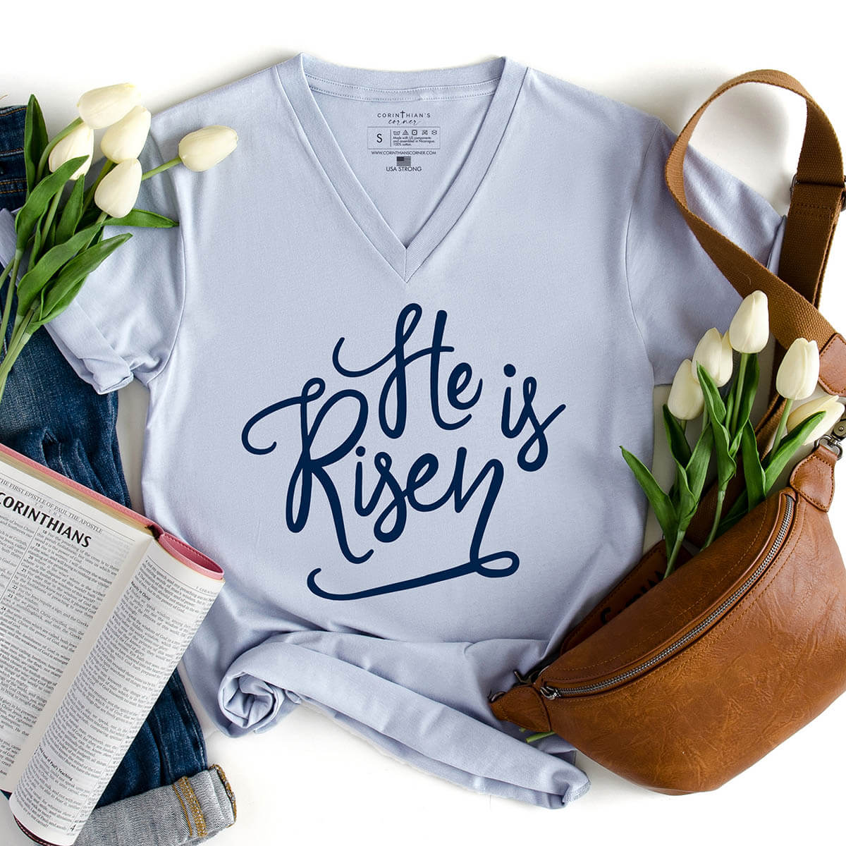 Light blue t-shirt with 'He is Risen' text, surrounded by tulips and a Bible on a white background