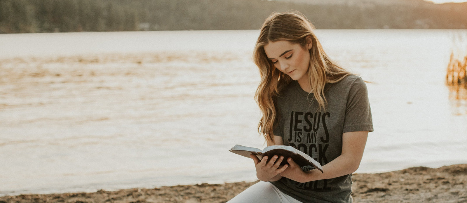 Woman reading a Bible by a lake in a Jesus Is My Rock shirt