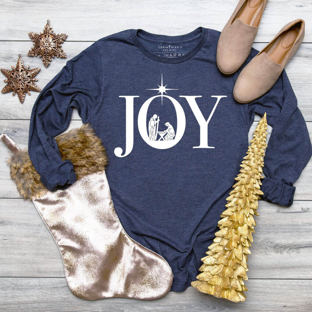 Uplifting manger scene displayed inside the word JOY printed on a premium long sleeve navy shirt