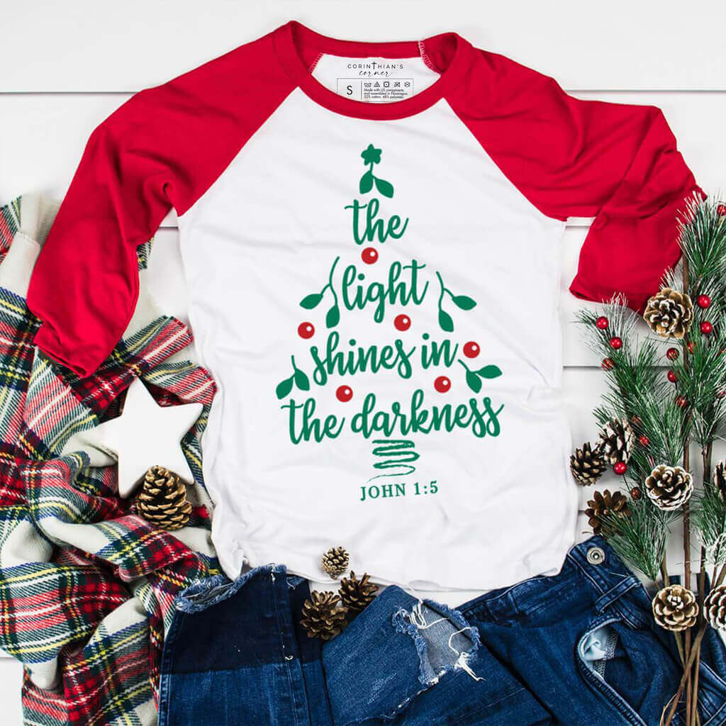 Christmas tree design in red and green that reads "the light shines in the darkness" on a red and white raglan shirt
