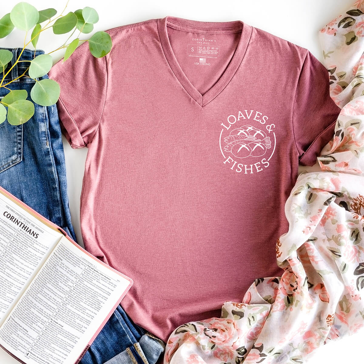 Pink t-shirt with 'Loaves & Fishes' design on a white surface with jeans, book, and plant.