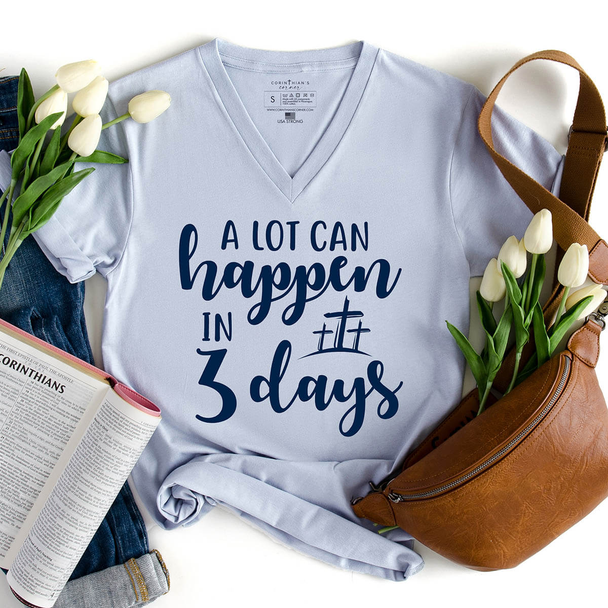 Light blue t-shirt with 'A lot can happen in 3 days' text, surrounded by flowers and a bag on a white background