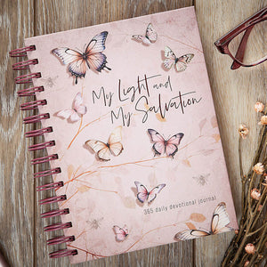 A spiral-bound devotional journal titled 'My Light and My Salvation' lies on a rustic wooden surface, surrounded by decorative props including eyeglasses and dried flowers. The cover is a soft blush pink with watercolor-style butterflies and delicate botanical line art.