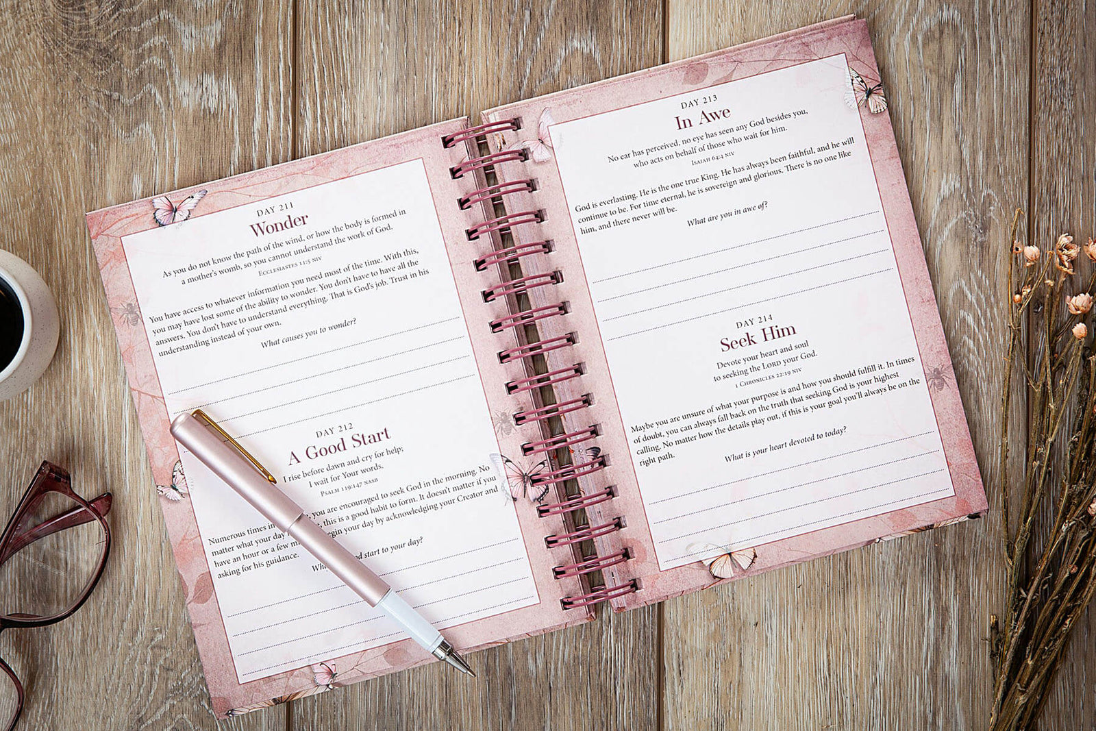 A spiral-bound devotional journal titled 'My Light and My Salvation' lies on a rustic wooden surface, surrounded by decorative props including eyeglasses and dried flowers. The cover is a soft blush pink with watercolor-style butterflies and delicate botanical line art.
