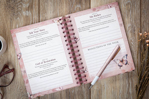 An open spiral-bound journal with pink floral pages laid on a wooden surface, alongside a rose-gold pen, eyeglasses, and dried flowers. The pages contain reflective journaling prompts such as 'Tell Your Story,' 'His Words,' and 'Reflections of the Week.