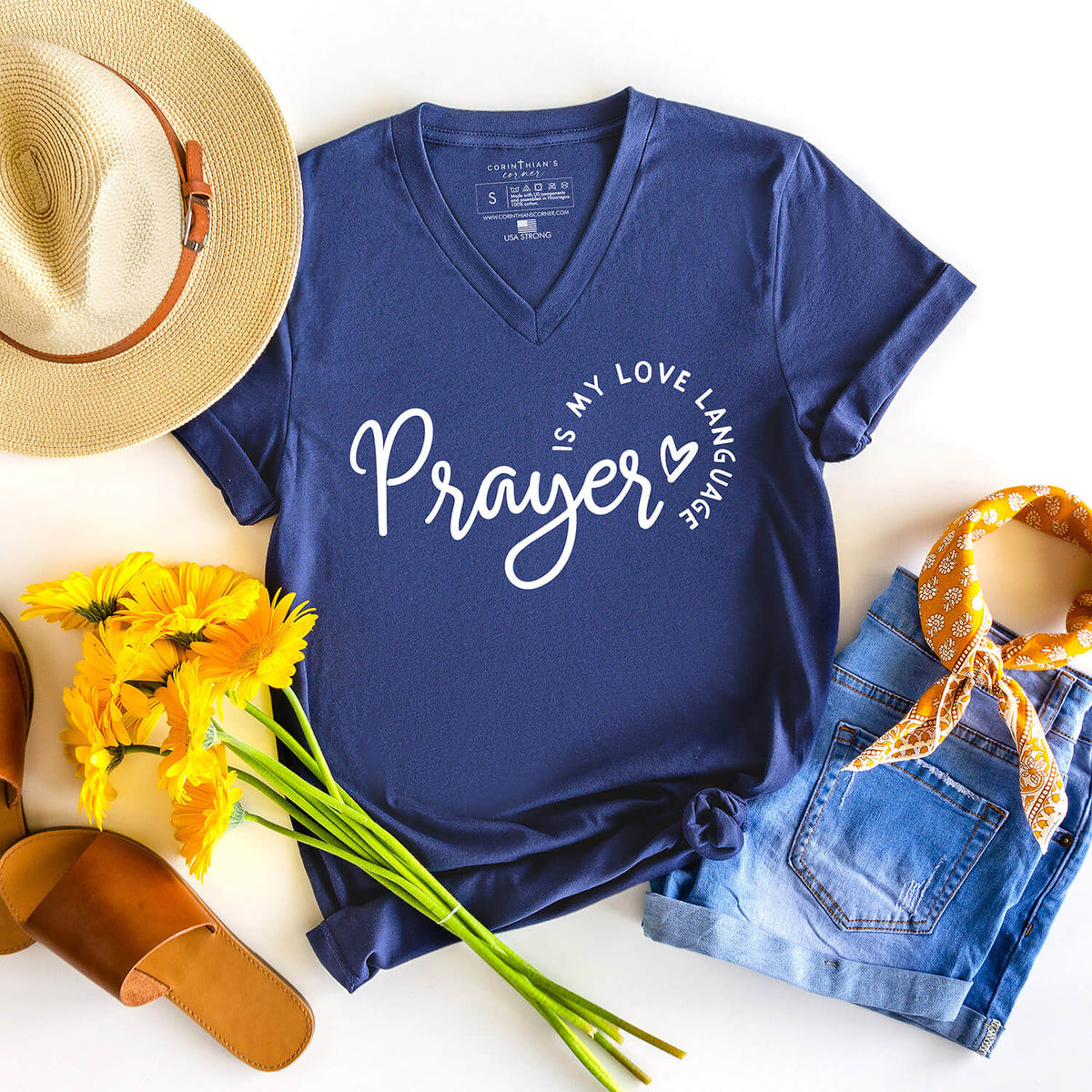 Navy blue t-shirt with 'Prayer is my love language' text, surrounded by a straw hat, yellow flowers, denim shorts, and sandals on a white background.
