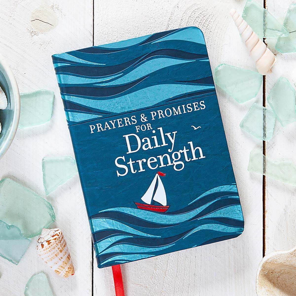 Book titled 'Prayers & Promises for Daily Strength' with a sailboat illustration on a wavy design, surrounded by seashells and glass stones.