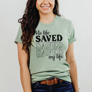 Smiling model against a plain background wearing the His life saved my life Romans 5:8 shirt