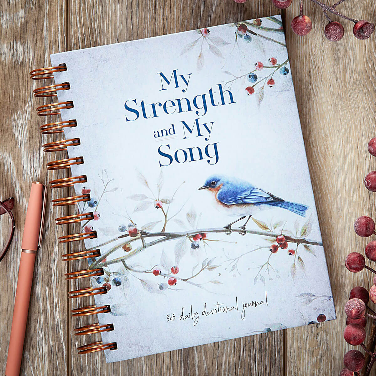 A spiral-bound devotional journal titled 'My Strength and My Song' lies open on a wooden surface, surrounded by decorative dried berries, a peach pen, and a pair of eyeglasses. The cover features a watercolor-style blue bird perched on a branch with small red berries on a pale background.