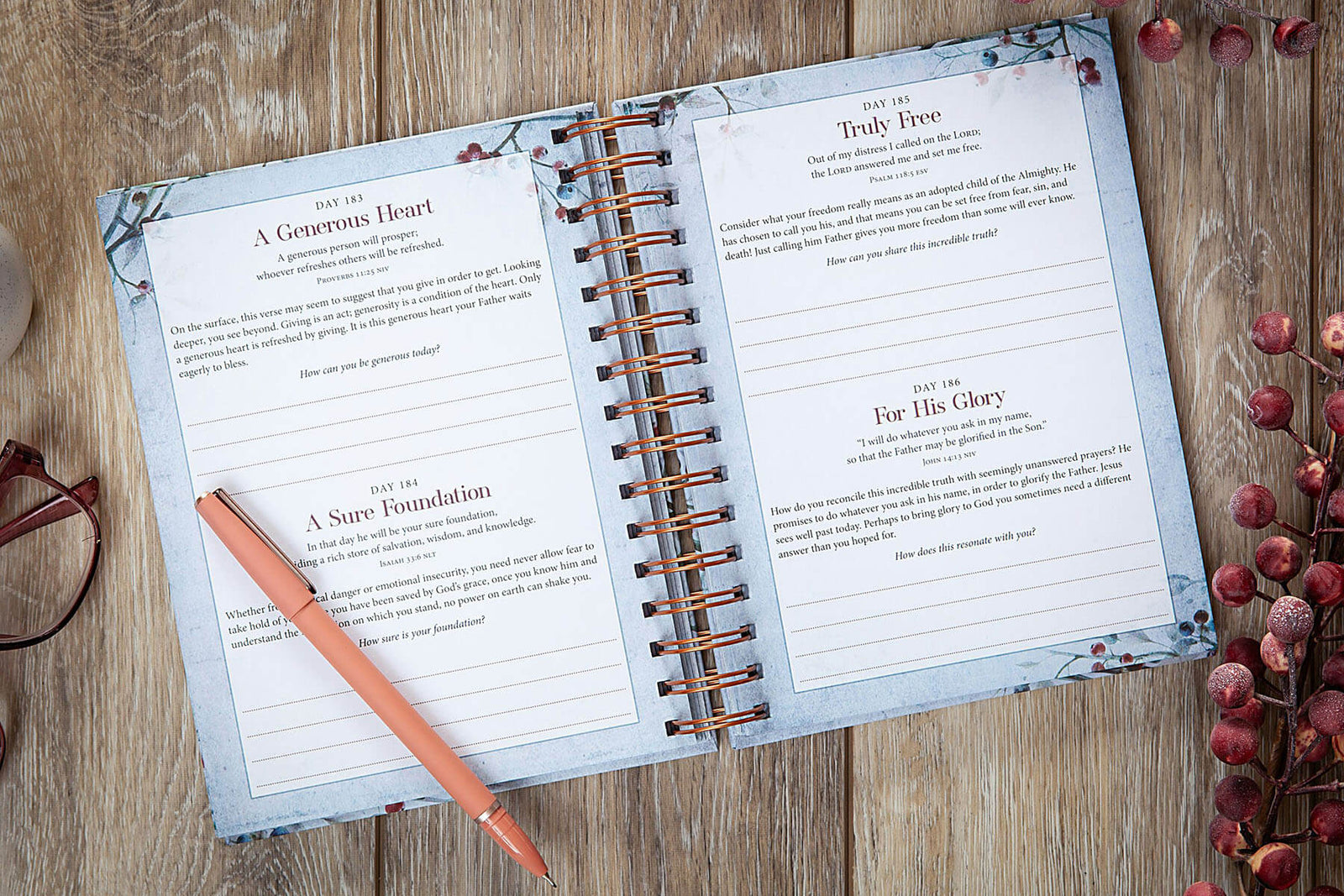 A spiral-bound devotional journal titled 'My Strength and My Song' lies open on a wooden surface, surrounded by decorative dried berries, a peach pen, and a pair of eyeglasses. The cover features a watercolor-style blue bird perched on a branch with small red berries on a pale background.