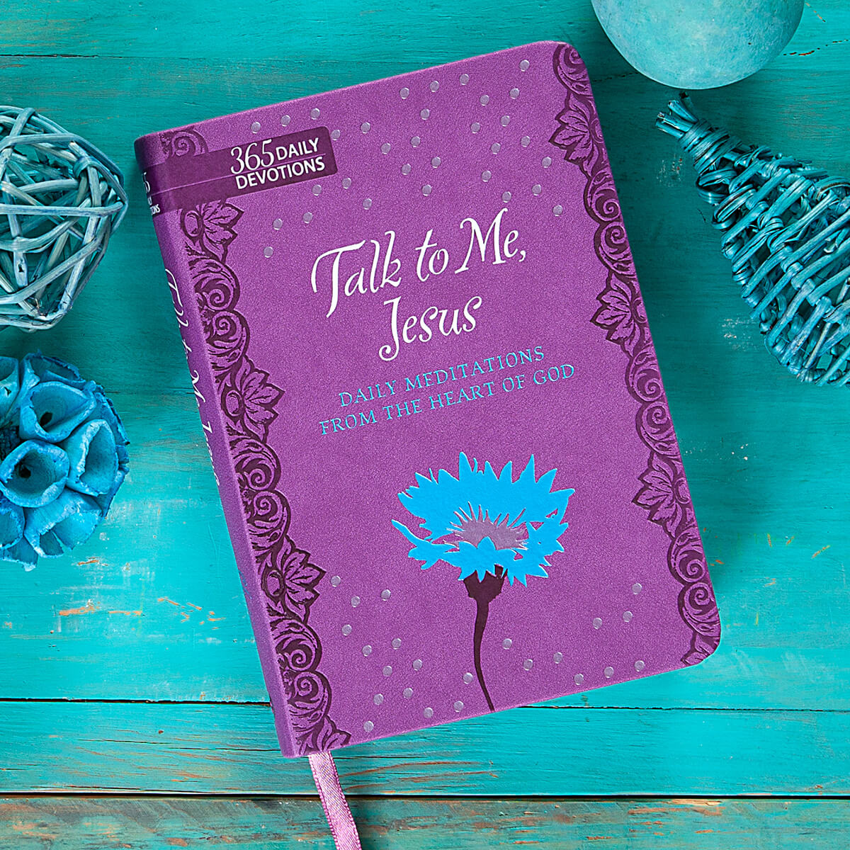 Purple devotional book titled 'Talk to Me, Jesus' on a teal wooden surface with decorative elements.