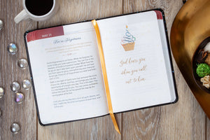 Open devotional with a decorative page on a wooden surface with a cup of coffee and decorative items.