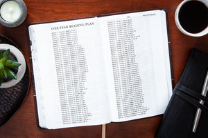 Open Bible with a one-year reading plan on a wooden table with a cup of coffee and candle.