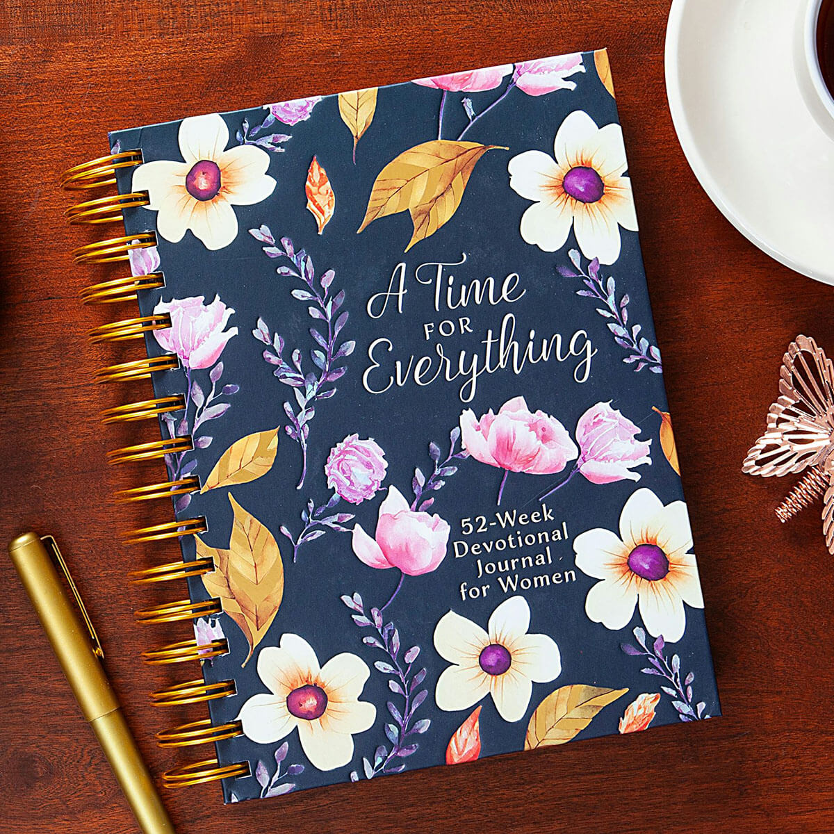 A floral spiral-bound devotional journal titled 'A Time For Everything' lies on a wooden table, styled with a gold pen, a white coffee cup, and a rose gold decorative object.