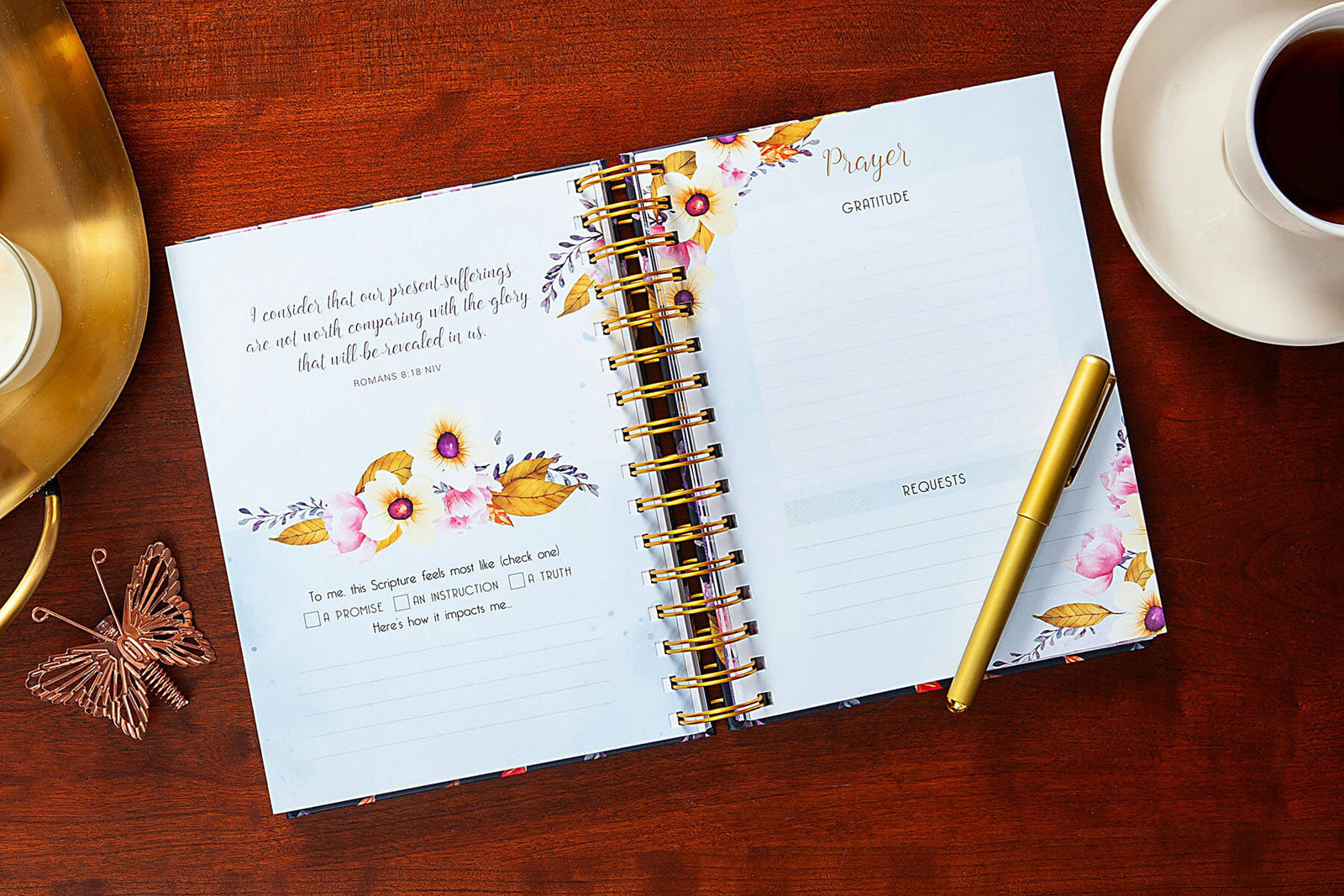 A floral spiral-bound devotional journal titled 'A Time For Everything' lies on a wooden table, styled with a gold pen, a white coffee cup, and a rose gold decorative object.