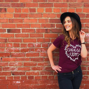 Woman smiling and leaning against a brick wall wearing choose joy Christian shirt